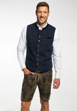 Dunkelblauer Weste mit silbernen Knöpfen und einer samtigen Tasche, kombiniert mit braunen Lederhosen mit geschnitzten Designs und leichten Stickakzenten.