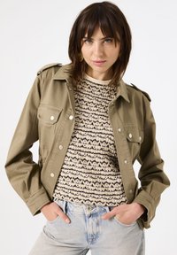 Olijfgroene katoenen jas met zilveren knopen, epauletten op de schouders en twee borstzakken, gedragen over een gestructureerde beige en zwarte trui.