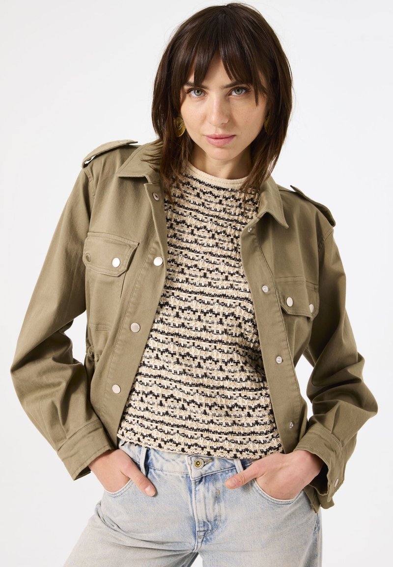Olijfgroene katoenen jas met zilveren knopen, epauletten op de schouders en twee borstzakken, gedragen over een gestructureerde beige en zwarte trui.