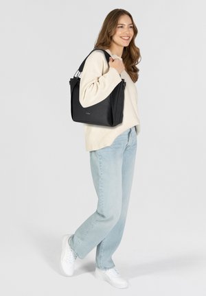 Sac fourre-tout en cuir noir avec finition texturée, doté d'une large ouverture et de doubles poignées, tenu par une personne portant un pull crème et un jean clair.