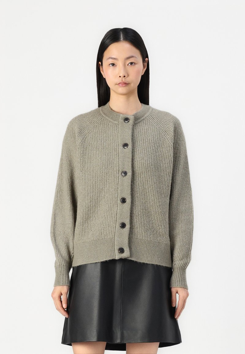 Strickjacke in hellem Olivgrün, mit einem gerippten Muster, rundem Ausschnitt und Knopfverschluss vorne. Kombiniert mit einem schwarzen Ledergemälde.