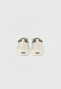 Hopeakosket sneakersit, joissa on teksturoitu pinta, kerma valkea pohja ja oranssit vetonauhat, joissa lukee "TINY TINY TINY", sekä brändäys kannassa.