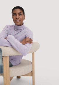 Lichtpaarse gebreide cardigan over een lavendel turtleneck, zittend op een afgeronde beige gestoffeerde stoel met houten poten, met casual denim.