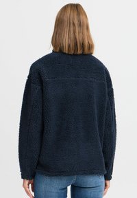 Veste en polaire bleu marine avec une surface texturée, dotée d'un col haut et de manches tombantes, portée sur un jean bleu clair.