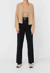 Beige, flauschiger Cardigan mit Knopfdetails, getragen über einem passenden Strickoberteil, kombiniert mit lockeren schwarzen Hosen und beigen Turnschuhen.
