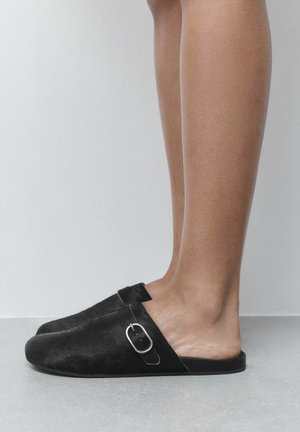 Person wearing black slip-on shoes with silver buckles, standing on a gray floor against a white wall background.