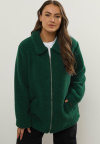 Zielona polarowa kurtka z zamkiem z przodu, oversize'owy kołnierz, dwie boczne kieszenie i teksturowany materiał. Nosić na białej koszuli i czarnych spodniach.