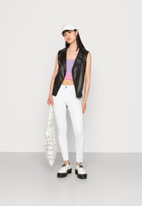 Jaqueta sem mangas de couro preto sobre um top cropped estampado rosa, combinada com jeans skinny brancos e sapatos brancos com cadarços pretos. A bolsa apresenta um padrão.