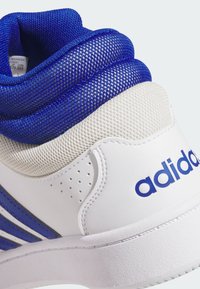 Kratki kadar bijele i plave Adidas tenisice s naglaskom na petu, koja pokazuje mrežastu tkaninu i Adidas logo sa strane.