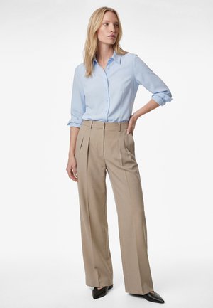 Donna con capelli biondi che indossa una camicia azzurra con bottoni, pantaloni beige a vita alta e gamba larga, e scarpe nere con punta, in piedi con una mano sul fianco.