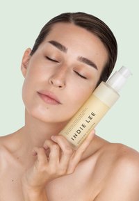 Oczyszczający kosmetyk INDIE LEE w przezroczystej, jasnobeżowej butelce z białą pompką, trzymany na nagim ramieniu. Delikatne, neutralne tło.