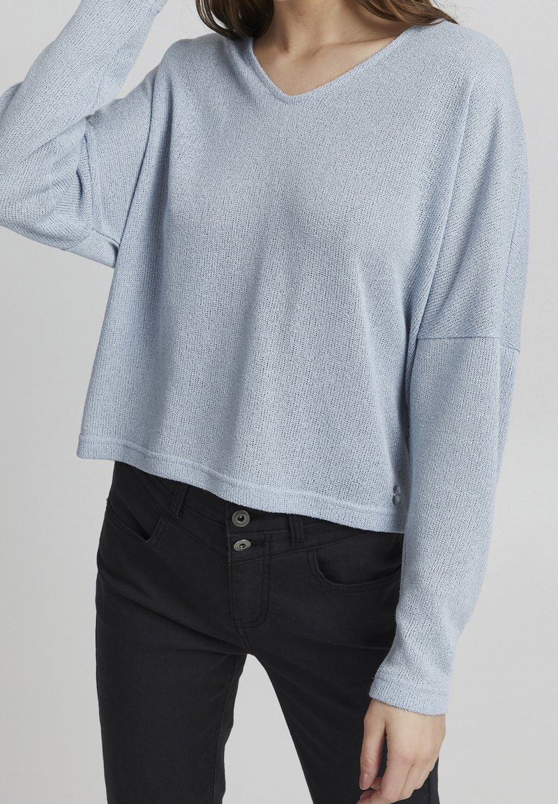 Lichtblauwe, gestructureerde gebreide trui met een V-hals en een drop-shoulder ontwerp. Gecombineerd met zwarte broek met twee metalen knopen.