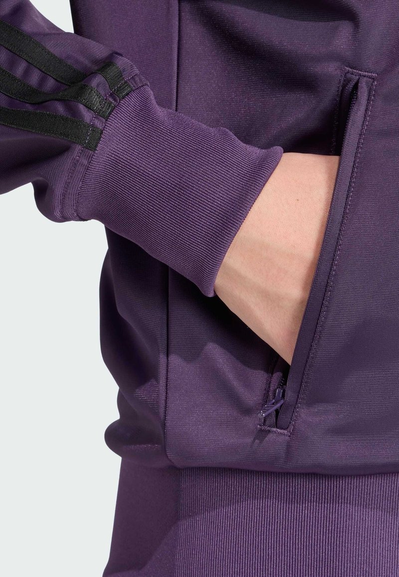 Giacca sportiva viola con strisce nere, tasca con cerniera, polsini a costine e texture liscia, progettata per comfort e facilità di movimento.