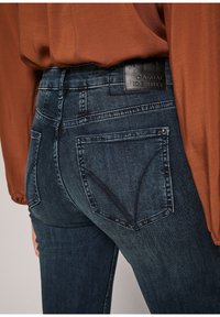 Gros plan d'une personne portant un jean bleu foncé avec des poches arrière et une étiquette « CASUAL IDENTITY », associé à un haut manches longues couleur rouille.