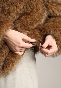 Kéz, amely egy barna, szőrös kabát húzókáját igazítja egy világos bézs szoknya vagy nadrág fölött, semleges háttér előtt.