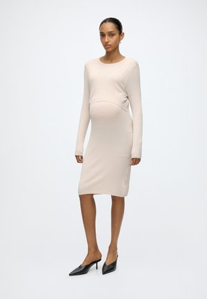 Femme aux cheveux foncés portant une robe midi beige côtelée à manches longues ajustée et des mules noires à talons hauts pointues, debout devant un fond uni.