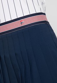 Marinblå plisserad kjol med ett vitt och marinblått randigt midjeband. Har en liten logga på midjebandet, tillverkad av mjukt, lätt tyg.