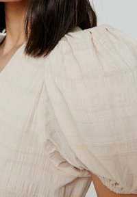 Blouse beige clair en tissu texturé froissé. Présente des manches bouffantes avec des détails froncés et des accents rayés subtils.