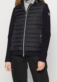Gilet imbottito nero con cuciture orizzontali e colletto alto, chiusura con zip argentata e tasche laterali. Maniche lunghe in tessuto uniforme.