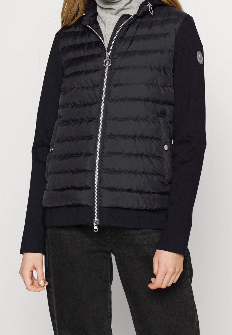 Gilet imbottito nero con cuciture orizzontali e colletto alto, chiusura con zip argentata e tasche laterali. Maniche lunghe in tessuto uniforme.
