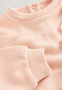 Sudadera rosa claro hecha de un tejido texturizado, con un cuello y puños acanalados, y un botón dorado decorativo en la parte posterior.