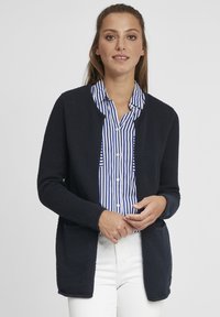 Cardigan texturé marine à ouverture sur le devant, manches longues et poches. Porté par-dessus une chemise à boutons rayée bleu et blanc, associé à un pantalon blanc.