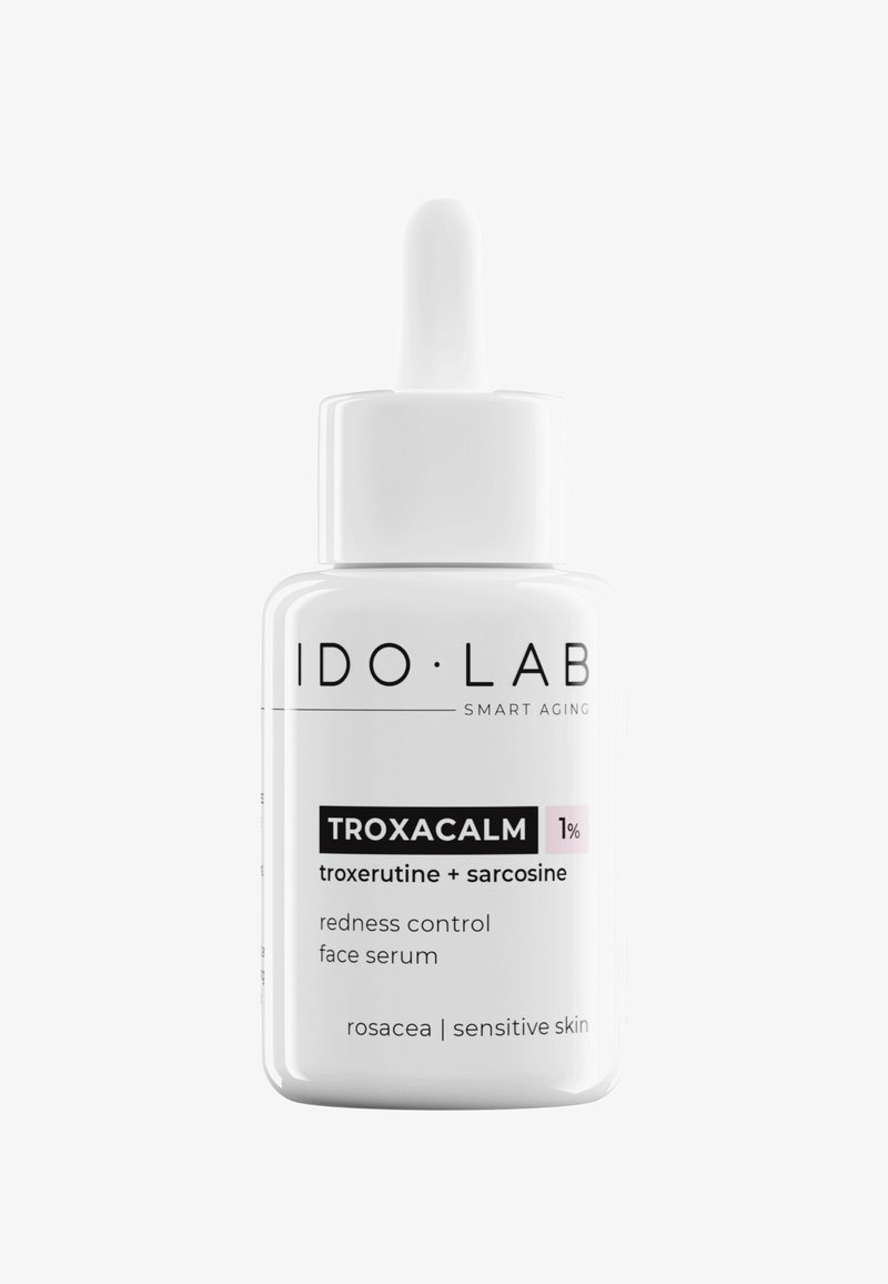 Biała szklana butelka z kroplomierzem, oznaczona etykietą "IDO•LAB TROXACALM 1%", zawierająca przezroczysty serum do twarzy kontrolujące rumień.