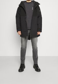 Schwarze Parkajacke mit Kapuze, ausgestattet mit einem Reißverschluss, zwei Farbdesign und verstellbaren Kordeln, kombiniert mit grauen Jeans und schwarzen Stiefeln.