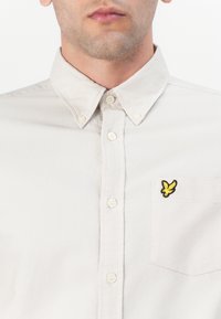 Svetlo bež srajca z gumbi in mehko teksturo, ki ima žep na levi strani prsnega dela in rumen vezen logotip ptice.