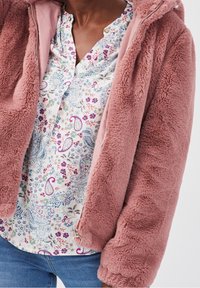 Person iført en delvist åbnet mauve fuzzy jakke over en blomster paisley mønstret bluse og blå jeans, der er cropped ved taljen.