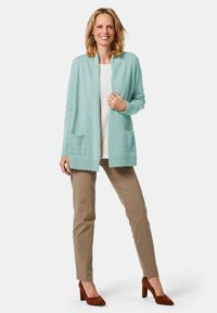 Hellblauer Strickcardigan mit offener Vorderseite, gerippten Bündchen und zwei aufgesetzten Taschen, kombiniert mit beigen Hosen und braunen Pumps.