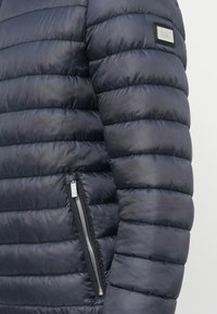 Detailný záber na rukáv námornícky modrej prešívanej páperovej bundy s vreckom na zips a nášivkou značky na hornej časti ramena.