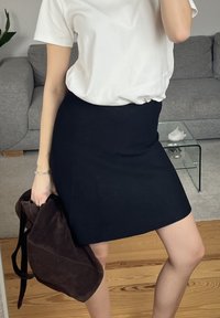 Kobieta w białej koszulce i czarnej spódnicy trzymająca ciemnobrązowy plecak, stojąca na drewnianej podłodze przed szarą sofą i szklanym stolikiem.