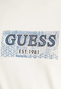 Camicetta bianca con una stampa grafica blu che mostra la parola "GUESS" e "EST. 1981" in grassetto nero, su uno sfondo testurizzato.