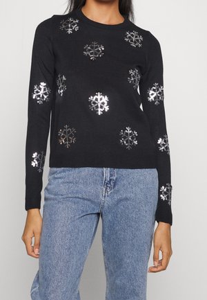 Pull noir en tricot avec des motifs de flocons de neige en paillettes argentées, col rond, manches longues et poignets côtelés. Associé avec un jean bleu clair.