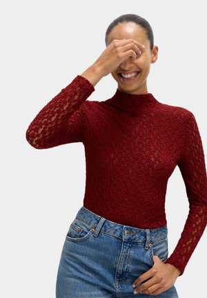 Top de encaje burdeos de manga larga con un cuello alto, que presenta una textura semitransparente, combinado con jeans de mezclilla de cintura alta.