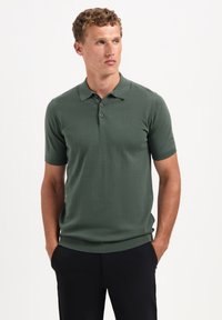 Groene polo met korte mouwen van zachte stof, met een drieknops sluiting en een geribde zoom. Gecombineerd met zwarte broek.