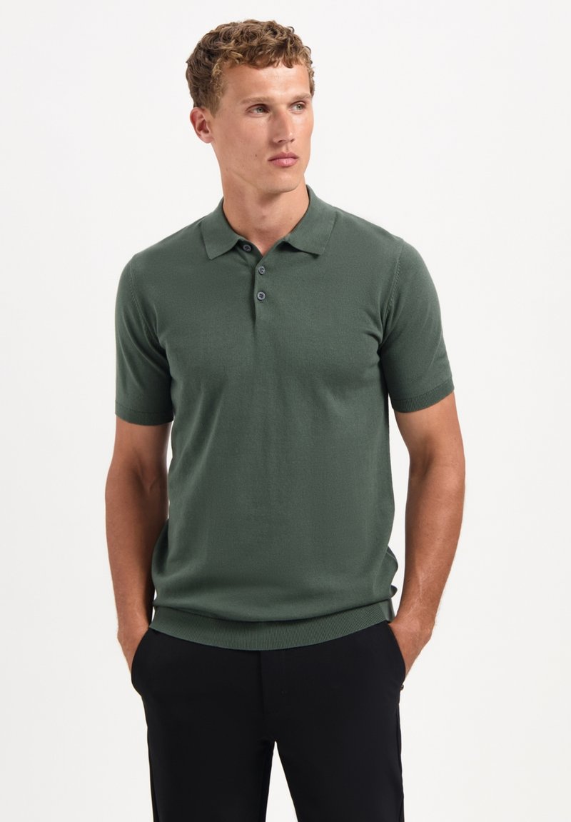 Groene polo met korte mouwen van zachte stof, met een drieknops sluiting en een geribde zoom. Gecombineerd met zwarte broek.