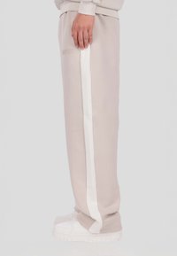 Bézs színű melegítőnadrág fehér oldalsávval, puha anyagból készült, dombornyomott logóval. Fehér tornacipővel párosítva.