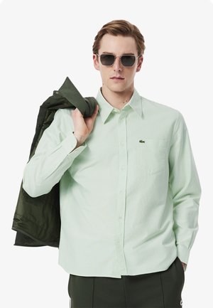 Camisa a rayas verde claro con cuello abotonado, que presenta un bolsillo en el pecho y un pequeño logo, sostenida con una chaqueta verde sobre el hombro.