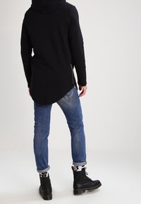Felpa nera lunga con orlo curvo, abbinata a jeans in denim blu e stivali neri con lacci. L'outfit presenta un look casual e stratificato.