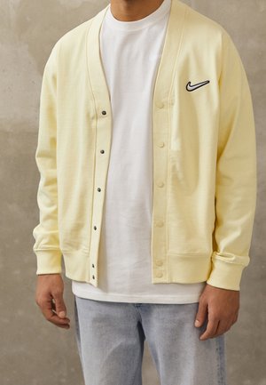 Mężczyzna ubrany w jasnożółty kardigan Nike na białej koszulce i jasnoniebieskich dżinsach, stojący na tle jednolitego szarego tła.