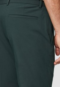 Dunkelgrüne Hosen mit glatter Textur und maßgeschneiderter Passform, versehen mit einer dezenten Gesäßtasche und Seitennähten für ein sauberes Design.