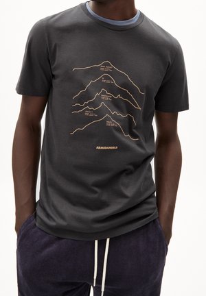 Personne portant un t-shirt gris foncé avec les contours et noms de montagnes, notamment le Mont Everest et le K2, assorti à un pantalon foncé avec des cordons blancs.
