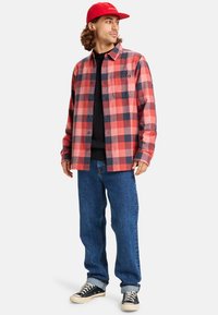 Rood en zwart geruite flanellen shirt met knopen, gedragen over een zwart topje, gecombineerd met blauwe spijkerbroek en zwarte sneakers, compleet gemaakt met een rode pet.