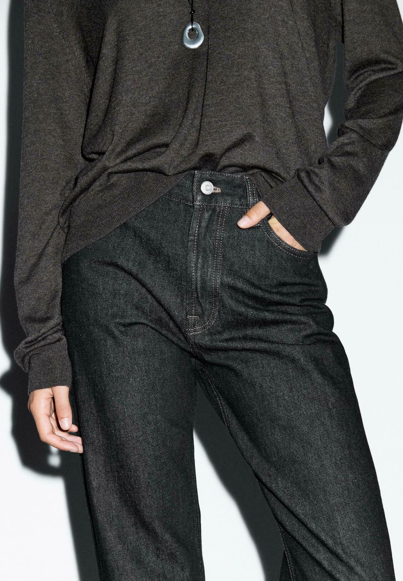 Tmavé denim kalhoty s vysokým pasem, viditelným švem a kapsami, v kombinaci s tmavým svetrem s dlouhým rukávem. Jednoduchý kulatý náhrdelník.