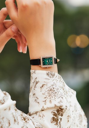 Reloj rectangular con una esfera verde, números romanos y acentos similares a diamantes sobre una correa de cuero negro, llevado en una mano. Tela con un patrón floral visible.