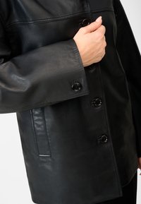 Sort læderjakke med en glat tekstur, der har store knapper, en brystseam detalje og en sidelomme.
