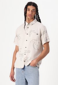 Camicia beige a maniche corte con disegni a linea nera di onde, palme e forme astratte; presenta una chiusura a bottoni e una tasca sul petto.