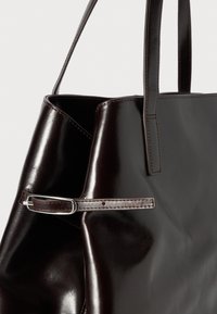 Dunkelbraune Ledertasche mit glatter Textur, schlanker Form und dünnem verstellbarem Riemen, der mit silbernem Zubehör gesichert ist.